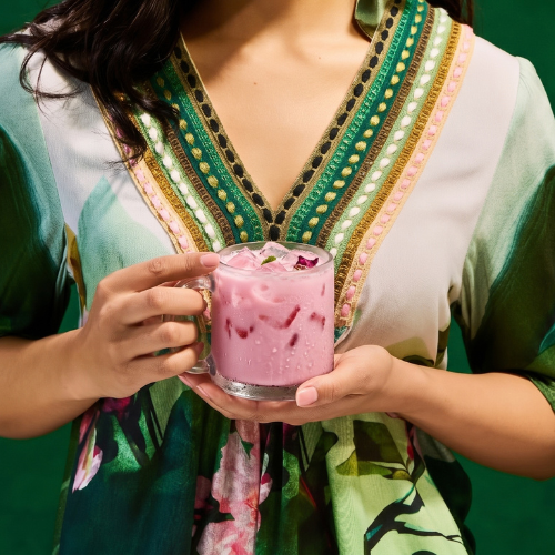 A person in a green and white patterned dress holds a glass mug filled with a pink iced drink against a solid green background.