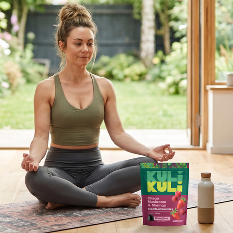Woman meditating cross-legged on a yoga mat at home beside a pouch of Kuli Kuli Chaga Mushroom & Moringa Superfood Gummies