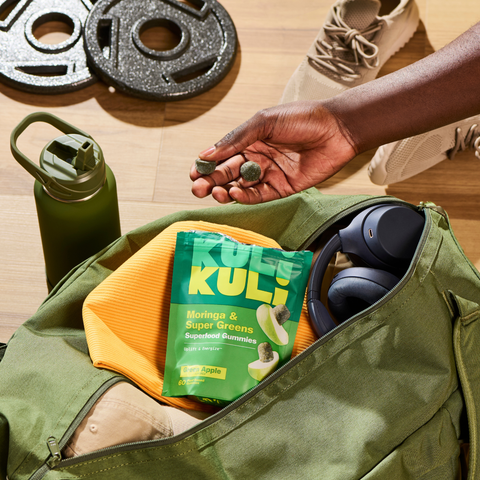 Hand holding two Kuli Kuli Moringa & Super Greens gummies above an open gym bag containing the Green Apple pouch, headphones, and a workout top, with weight plates, sneakers, and a water bottle nearby.