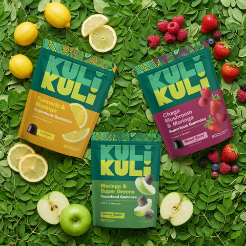 Three Kuli Kuli gummy pouches (Turmeric & Moringa, Chaga Mushroom & Moringa, and Moringa & Super Greens) laid on a bed of moringa leaves with lemons, mixed berries, and green apples.