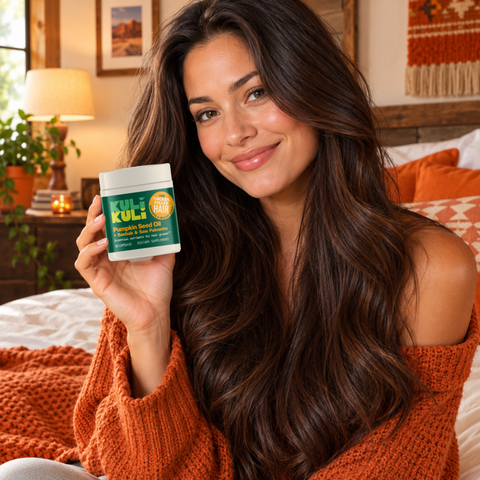 Smiling woman with long, glossy brown hair and an orange sweater holding a jar of Kuli Kuli Pumpkin Seed Oil + Baobab & Saw Palmetto capsules in a cozy bedroom.