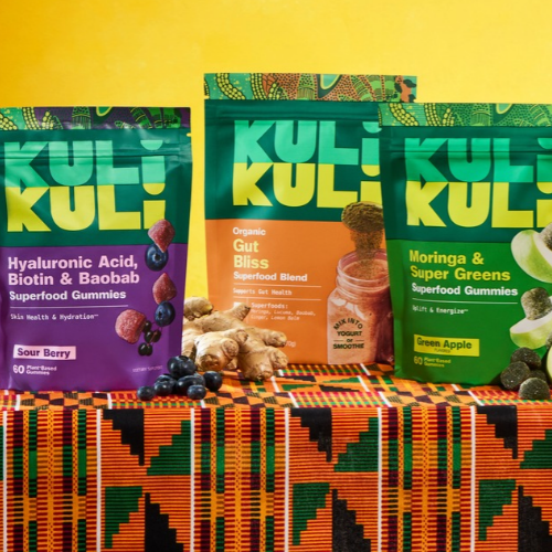 Three stand-up pouches of Kuli Kuli superfood gummies are displayed on a colorful patterned fabric against a bright yellow background. The pouches are for Sour Berry, Organic Gut Bliss, and Green Apple flavors, with ginger, blueberries, and fruit pieces arranged in front.