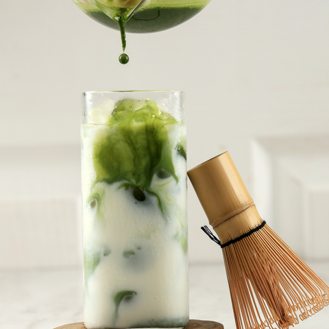 Bright green matcha pouring into a tall glass of iced milk, swirling as it blends, beside a bamboo matcha whisk.