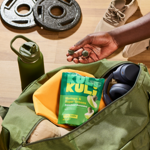 Person holding Kuli Kuli snacks next to a green bag with fitness equipment on a wooden floor.