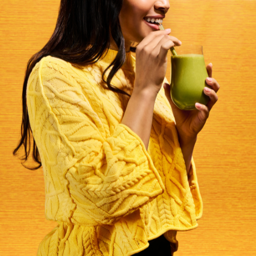A woman wearing a bright yellow cable-knit sweater drinks from a green glass with a straw, set against a warm yellow background.