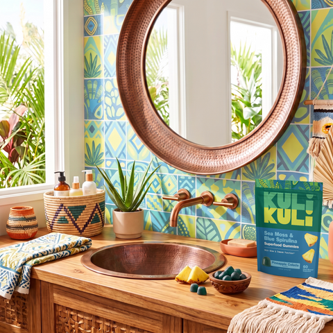 Kuli Kuli Sea Moss & Blue Spirulina gummies pouch on a bathroom vanity with a copper sink, colorful patterned tiles, aloe plant, pineapple slices, and a small bowl of gummies.
