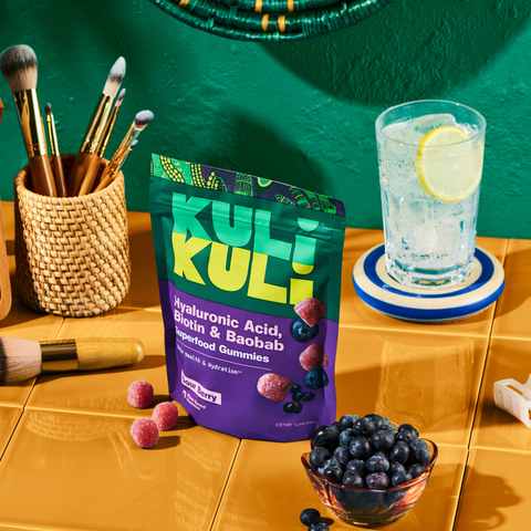 Kuli Kuli Sour Berry gummies pouch on a yellow tiled vanity beside makeup brushes, a glass of lemon water, a bowl of blueberries, and loose gummies.