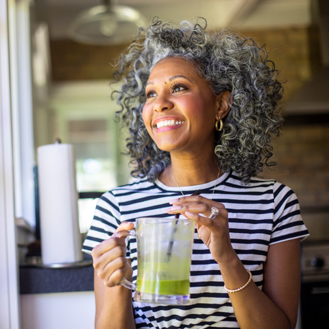 A smiling woman with curly gray hair holding a mug of green drink in a sunny kitchen.