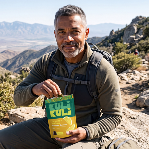 Smiling hiker with gray hair and a beard sitting on a mountain trail, taking a gummy from a bag of Kuli Kuli Turmeric & Moringa Gummies.